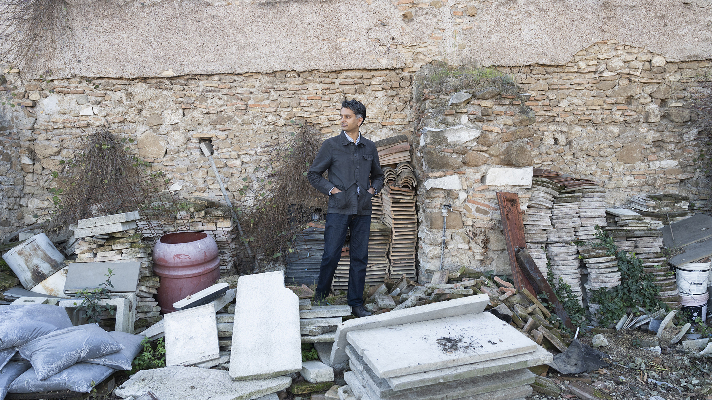 Color photo of a brown skinned man wearing a dark jacket and pants; he stands among strewn building construction material in concrete, terracotta, plastic, and sandbags