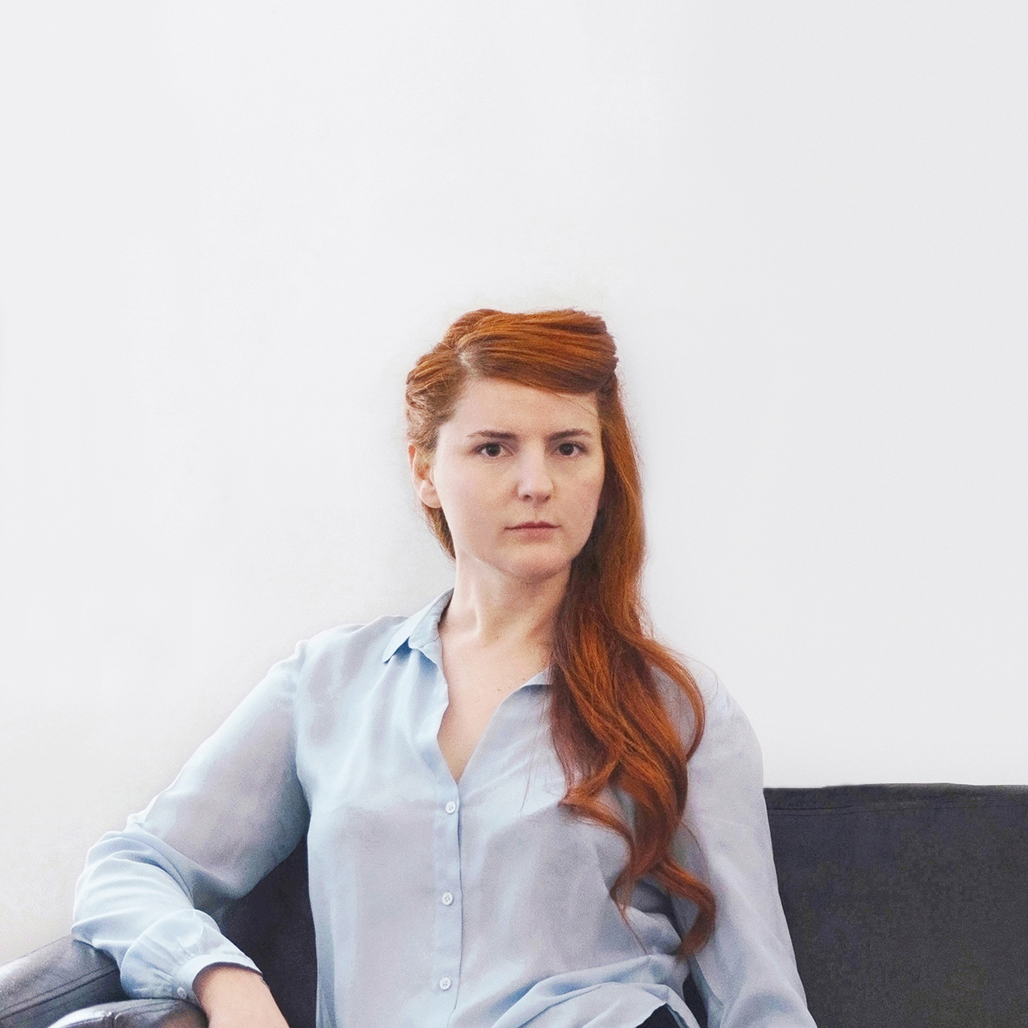 Color photograph of a light skinned woman with long red hair wearing a button-up shirt and looking at the camera