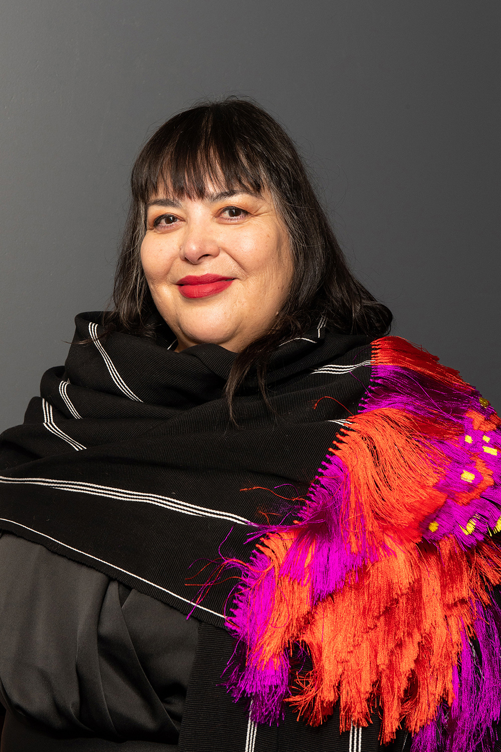 Color photograph of a light skinned woman with dark hair and bright red lipstick smiling at the camera; she wears a complex, multilayered garment in black, fucsia, and orange