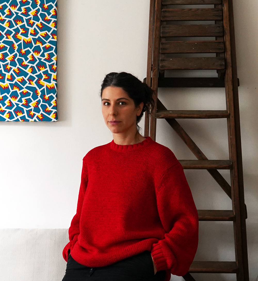 Color photograph of a light skinned woman wearing a red sweater; she stands indoors next to a ladder leaning on the wall, looking directly at the camera