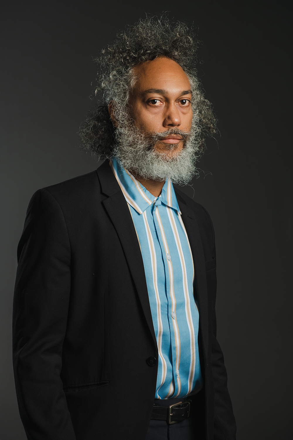 Color photograph of a dark skinned man with gray hair and beard wearing a dark blazer over a blue shirt; he looks at the camera with a serious expression