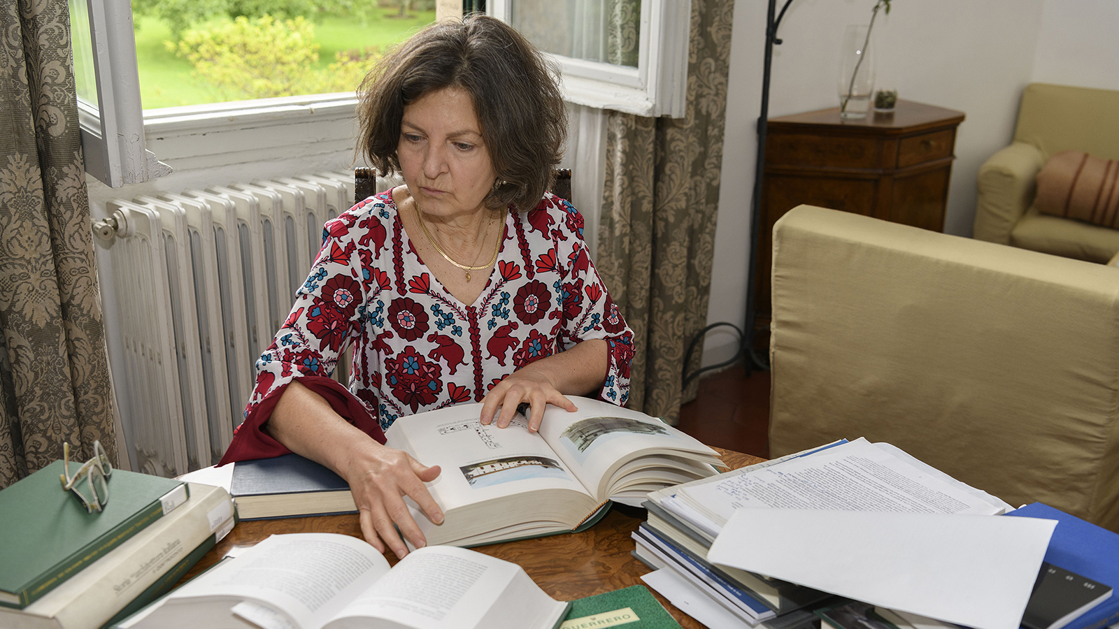 Color photograph of a light skinned woman seated at a desk covered with books, which she is poring over with her eyes and hands; behind her is an open window overlooking a garden
