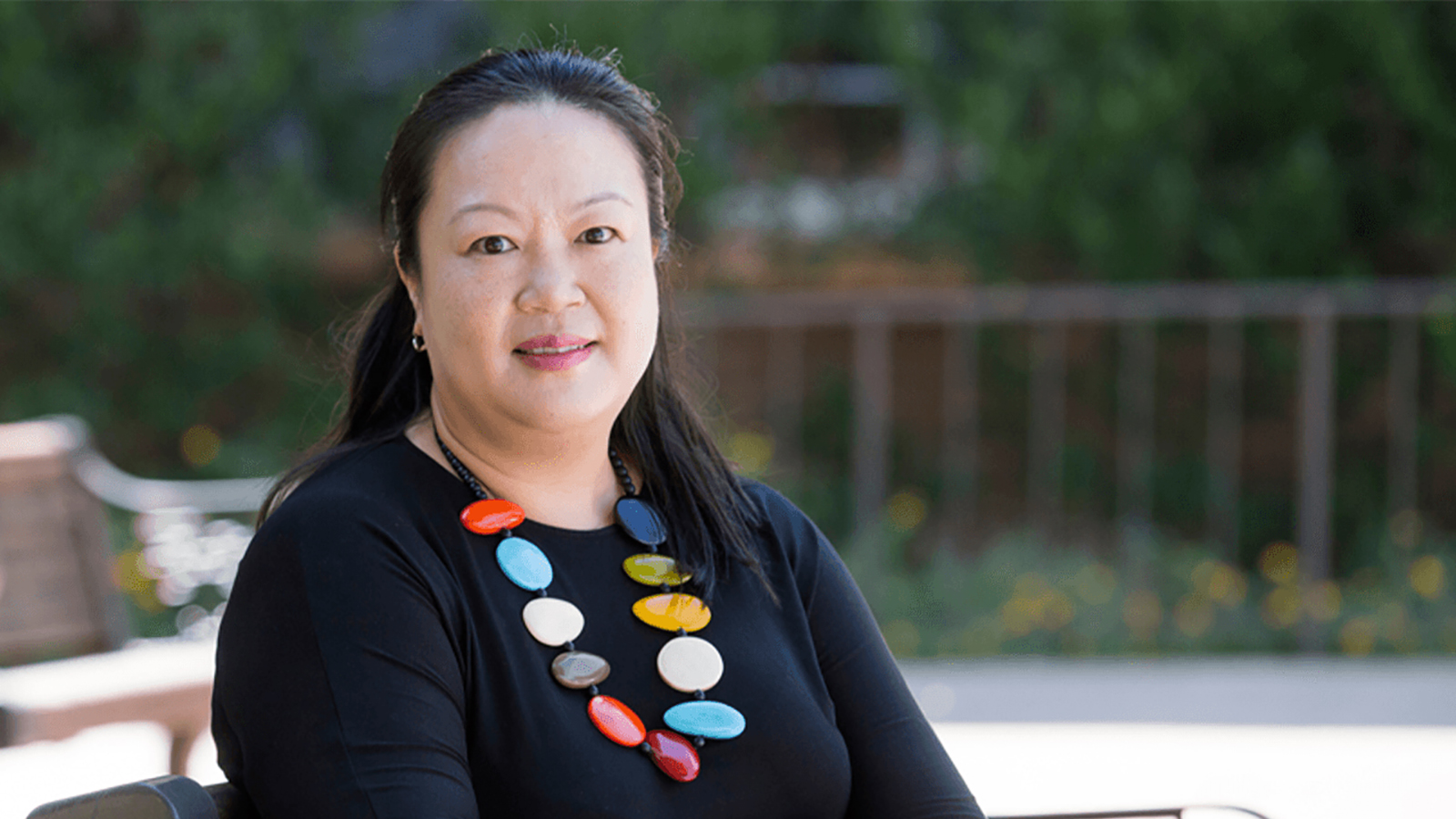 color photograph of an Asian woman sitting in an outdoor location and wearing a black top and a colorful painted wood or stone necklace; she smiles and looks directly at the camera