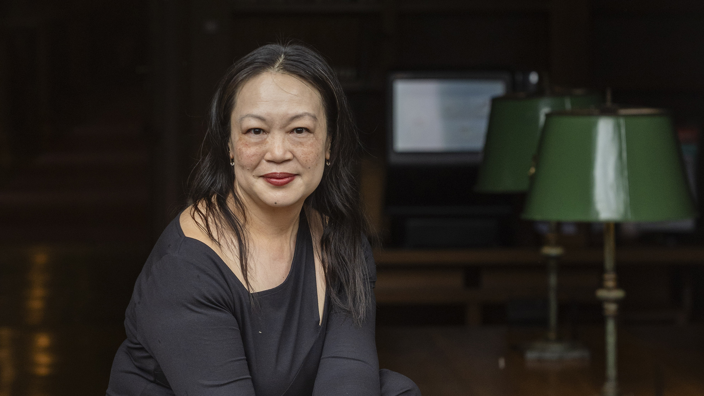 Color photograph of the head and shoulders of an asian woman with dark hair sitting at a library desk and looking at the camera