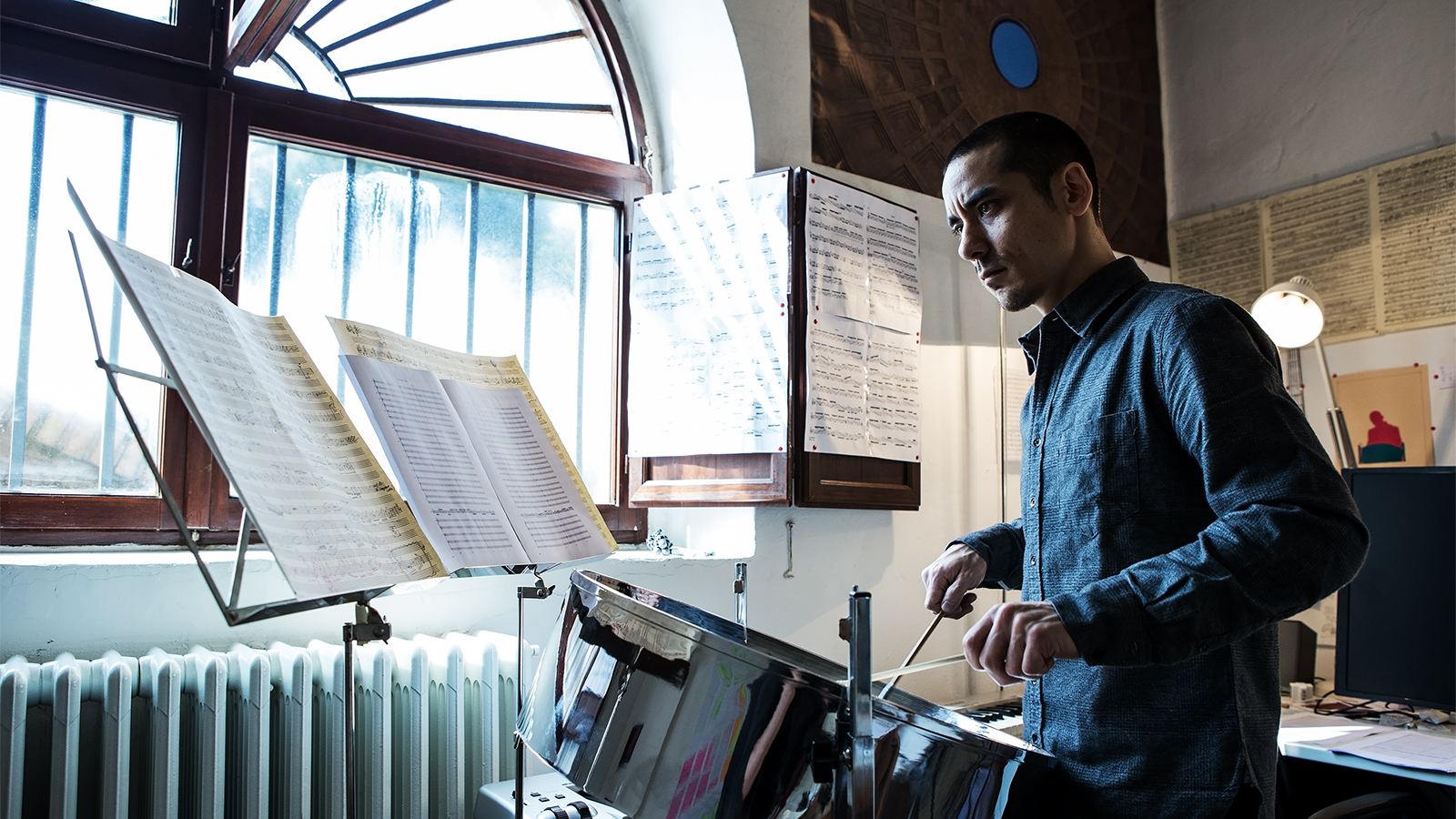 Color photograph of an Asian man playing a steel drum while looking at sheet music on a music stand