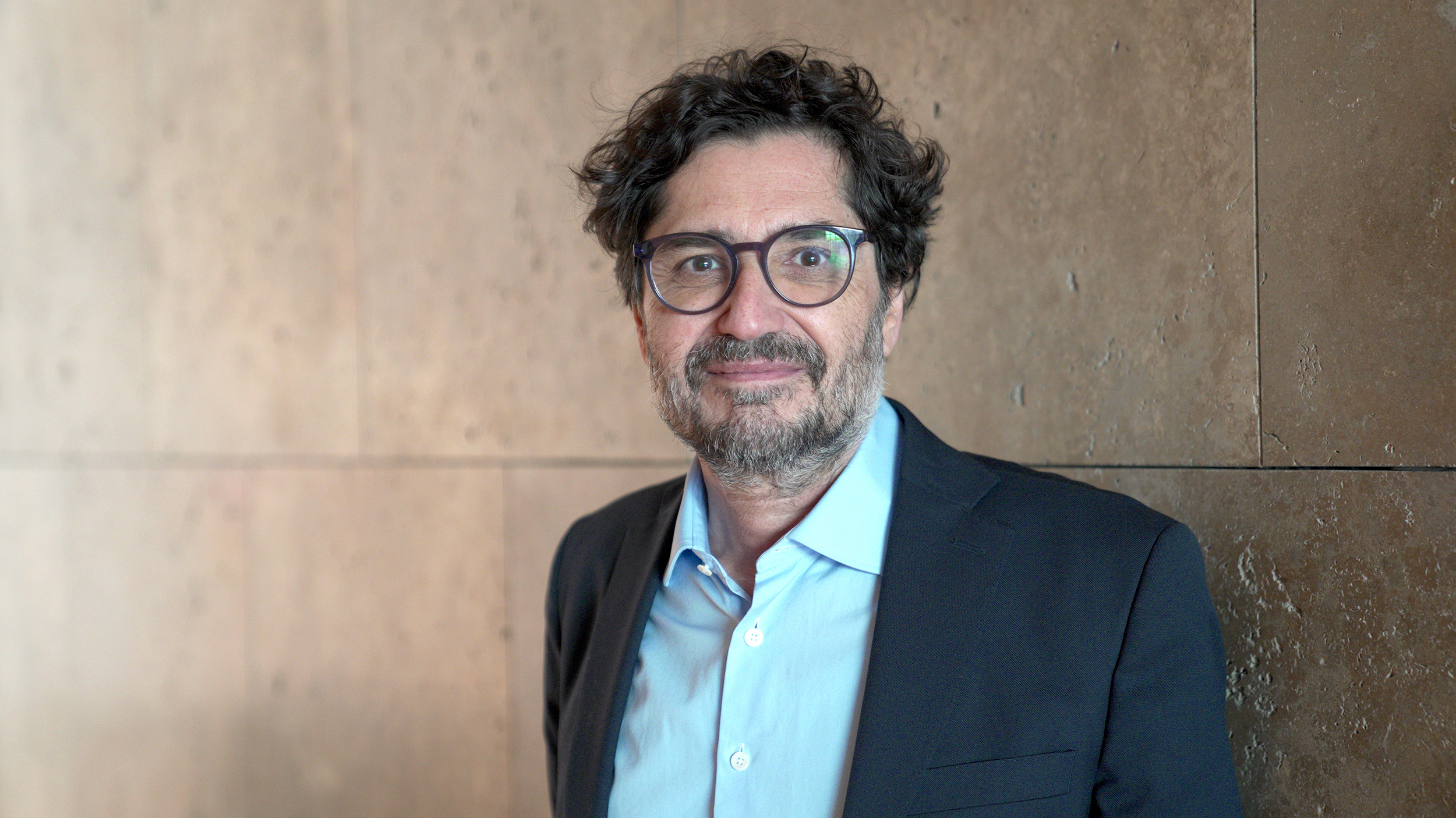 Color photograph of the head and torso of Avinoam Shalem, wearing a blue shirt and a charcoal blazer and standing against a sandy-colored wall