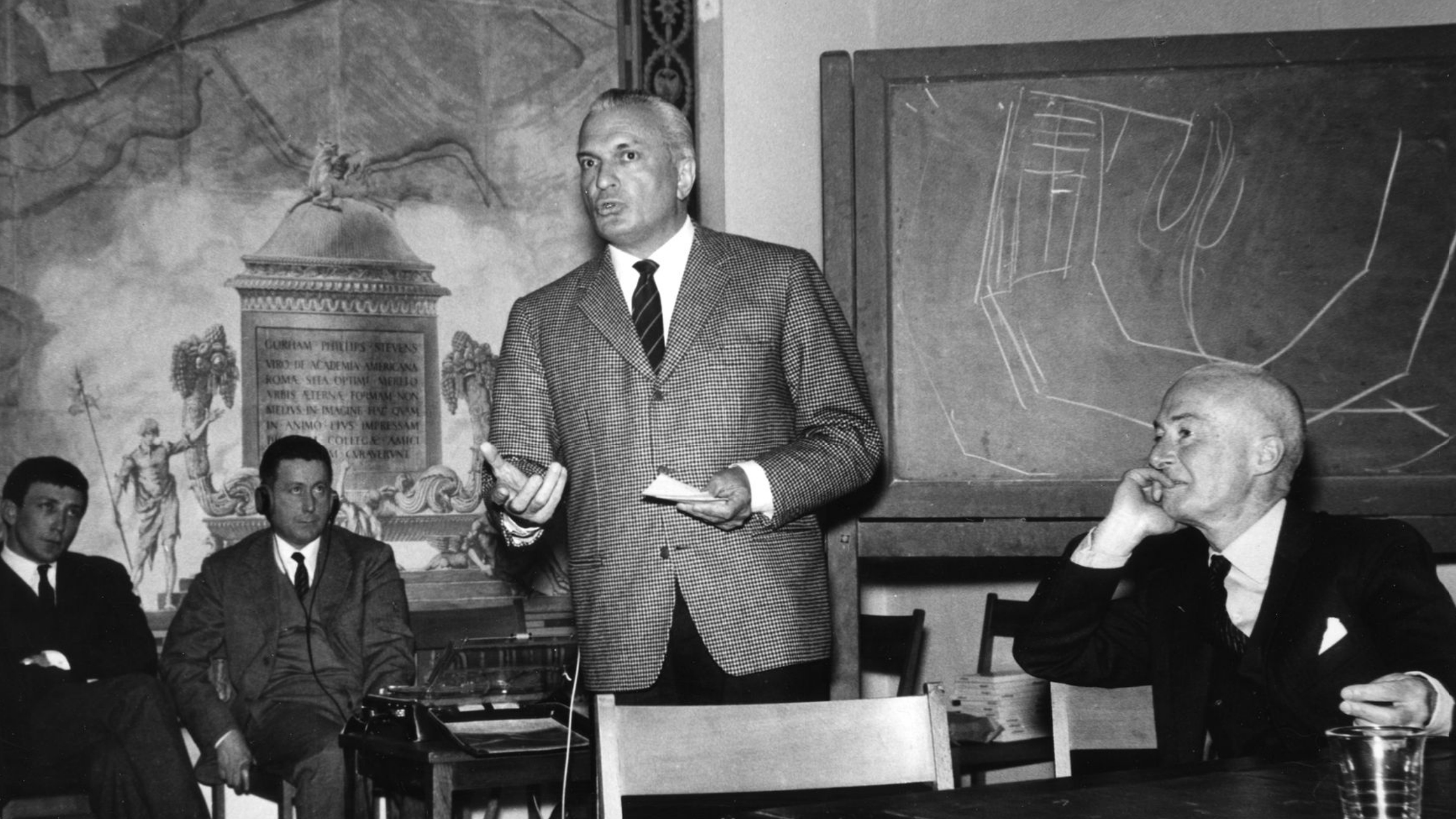 Black and white photograph of a light skinned man wearing a suit standing and speaking to various seated people at a conference