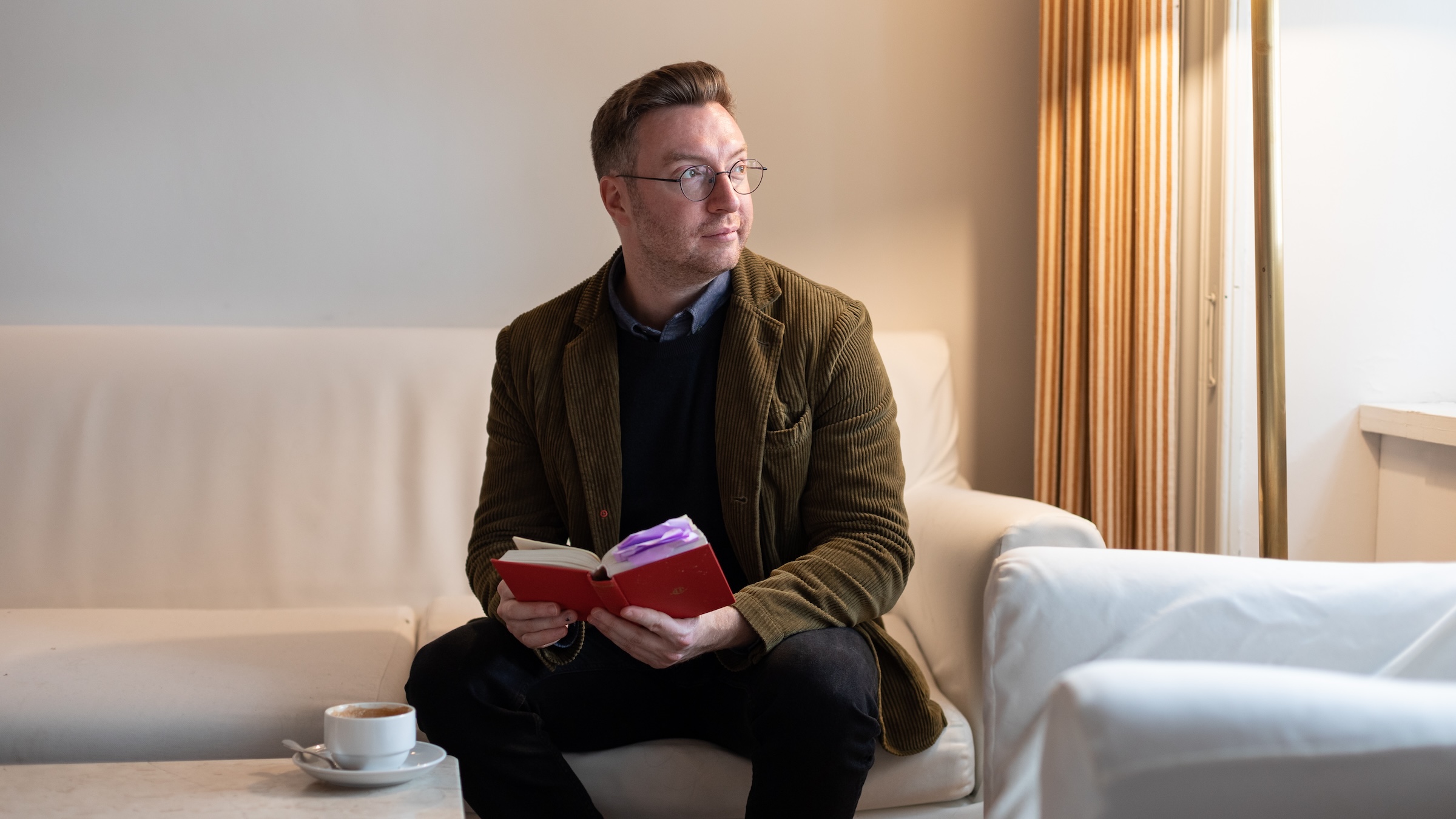 A young man holds a book with a red cover sitting on a white sofa, with light falling from the right