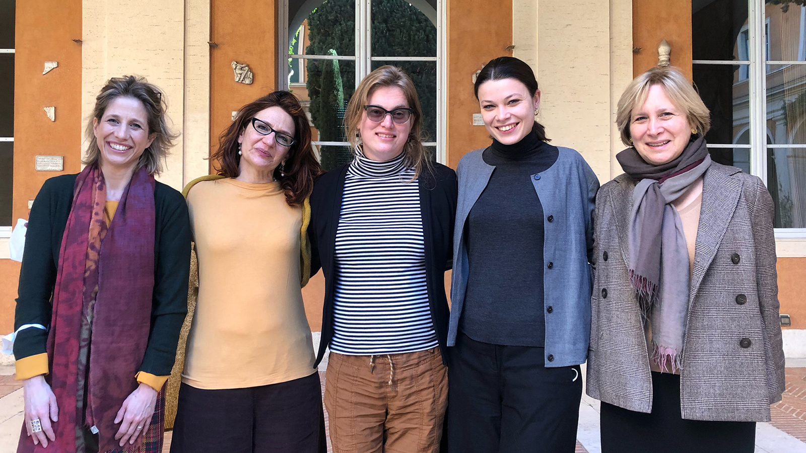 Color photograph of five light skinned women of varying ages standing in a row in an outdoor location, looking at the camera and smiling