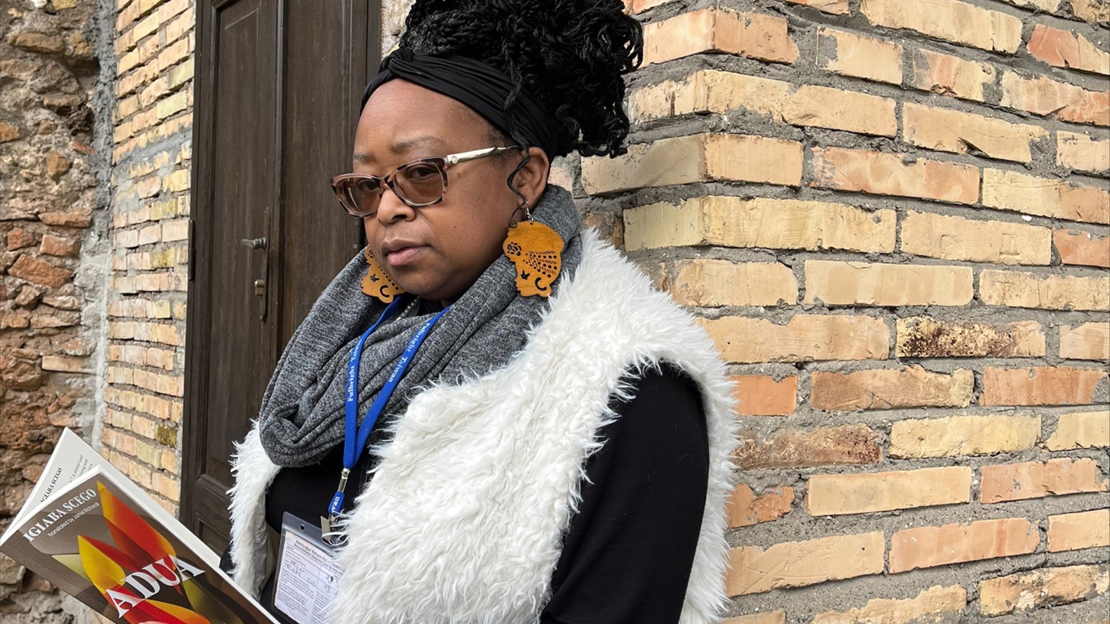 A darked skinned woman with dreadlocks and wearing sunglasses leans against a brick wall; she holds open a copy of Igiaba Scego's book Adua in her hands but looks at the camera