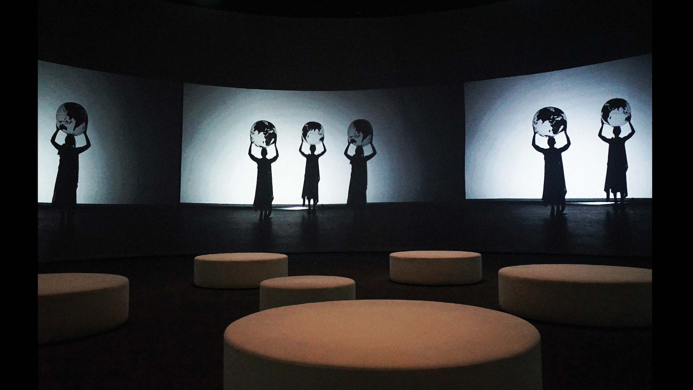 Color photograph of an indoor art installation showing a darkened room spotlit on the floor and with shadowy figures projected on the walls