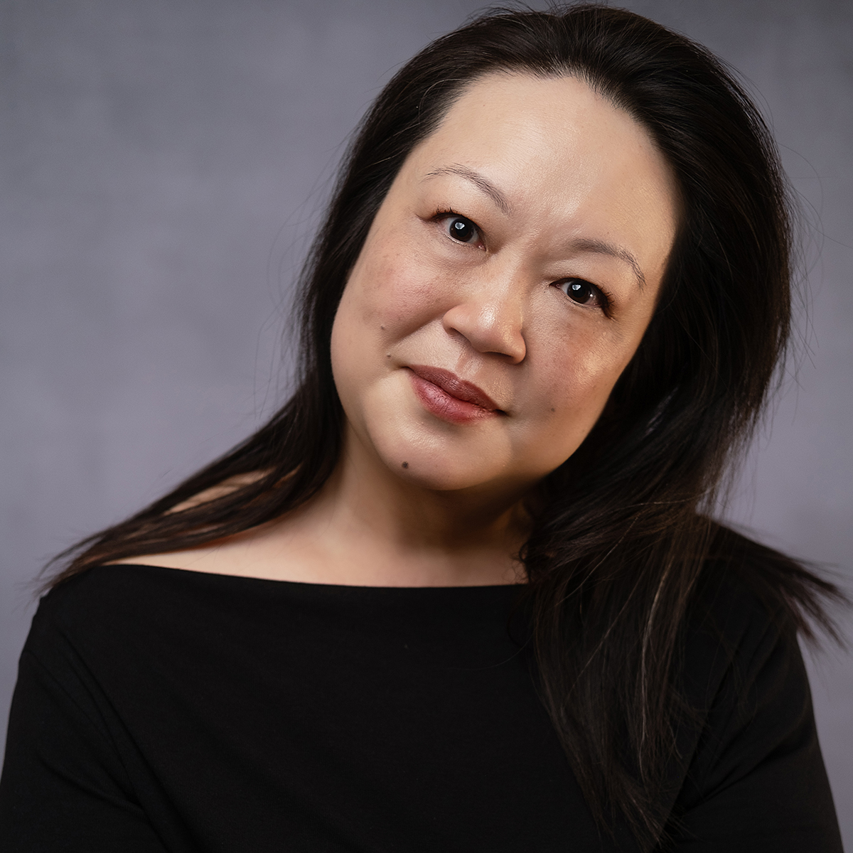 A color photograph of the head and torso of an Asian woman; she tilts her head to the left while looking directly at the camera