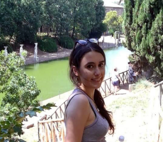 Arianna Brunori Color photograph of the head and shoulders of a light skinned woman with brown hair standing on a hill above a reflecting pool; she turns toward the camera and smiles