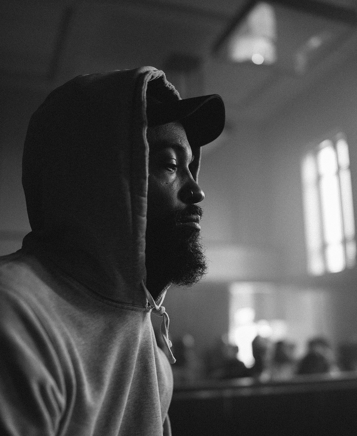 Bradford M. Young Black and white photograph of the head and shoulders of a dark skinned man with a beard and wearing a hoodie over a baseball cap; he is seen looking pensive in 3/4 view sitting in a high-ceiling room that has a blurry background