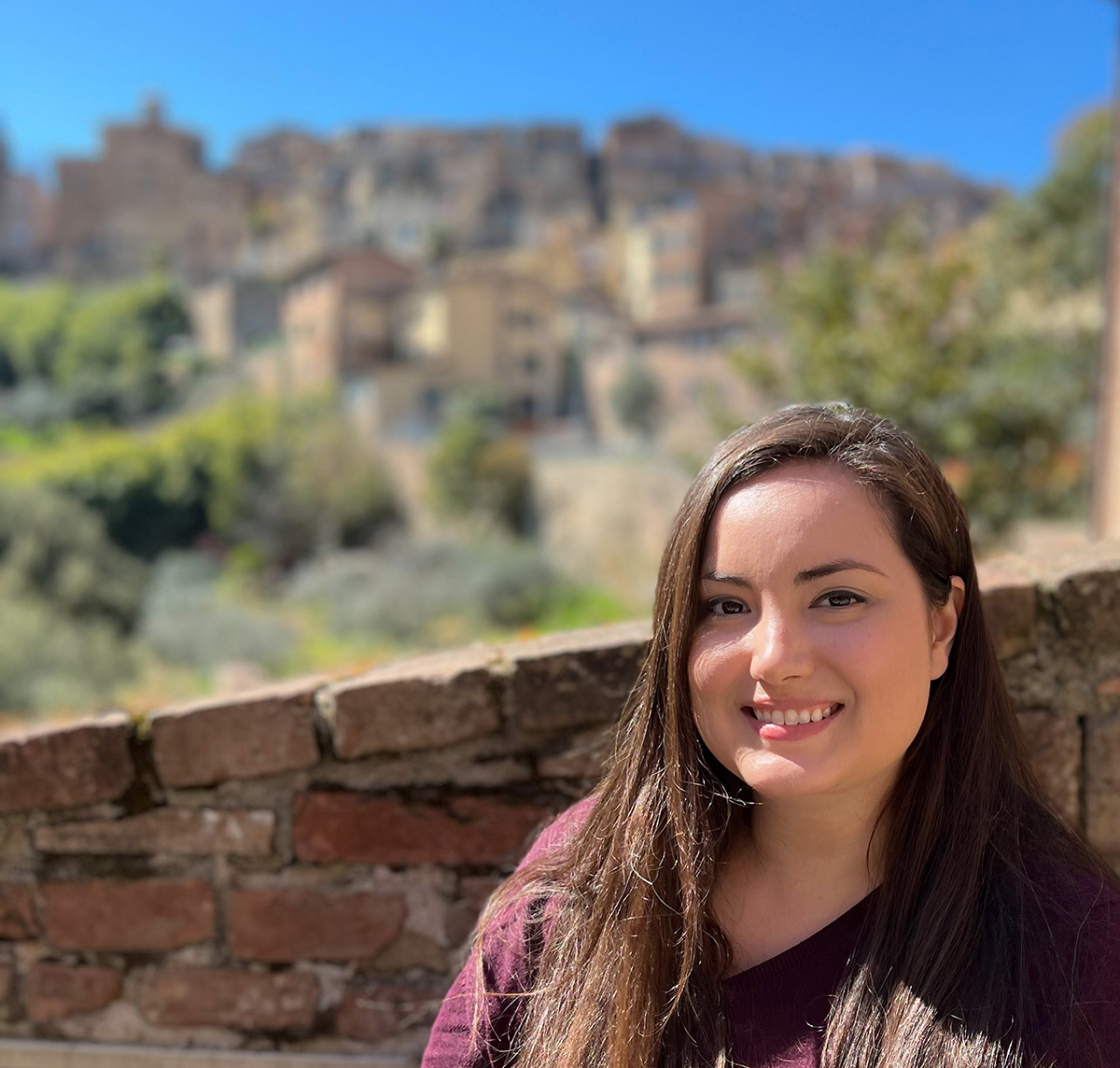 Carolyn J. Quijano Color photograph of the head and shoulders of a light skinned woman with long brown hair smiling at the camera; she is in an outdoor location, at a brick ledge overlooking a hillside with buildings