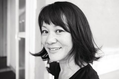 Black and white photograph of an Asian woman with straight dark hair looking at the camera over her shoulder and smiling