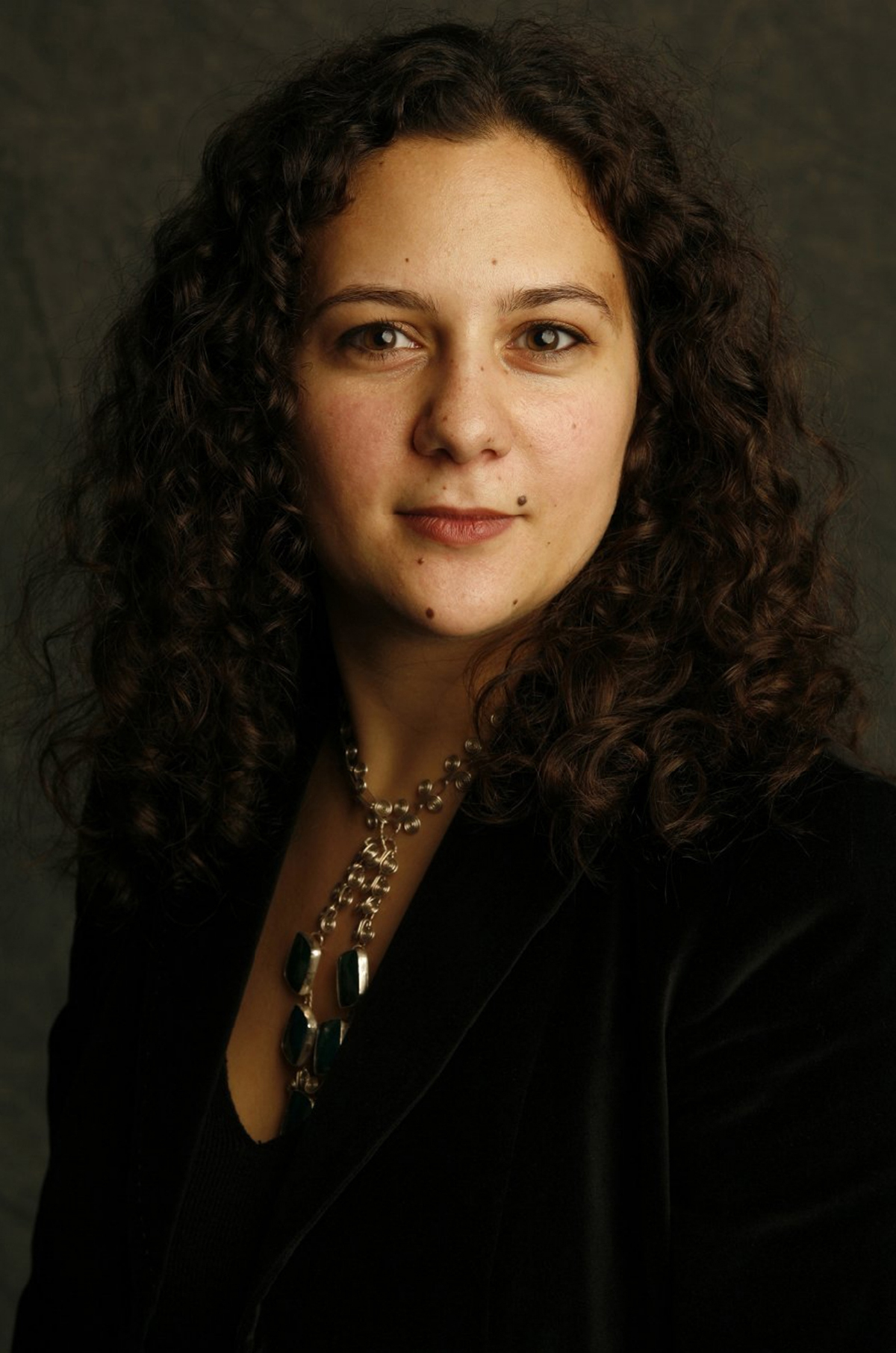 Christina Ferando Color photograph of the head and torso of a light skinned woman with dark hair and a silver necklace looking directly at the camera