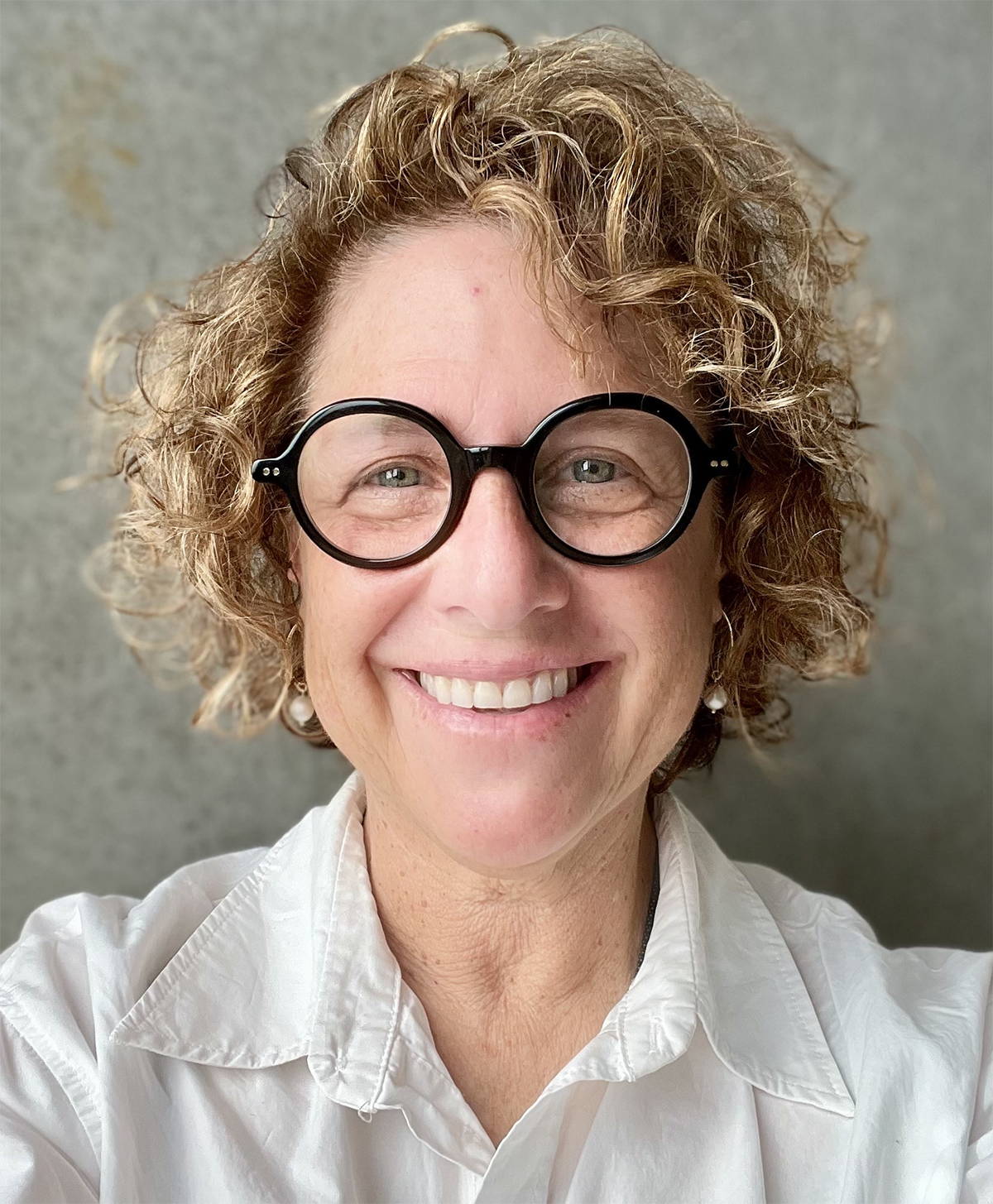Color photograph of a light skinned woman with curly hair and wearing glasses, smiling at the camera