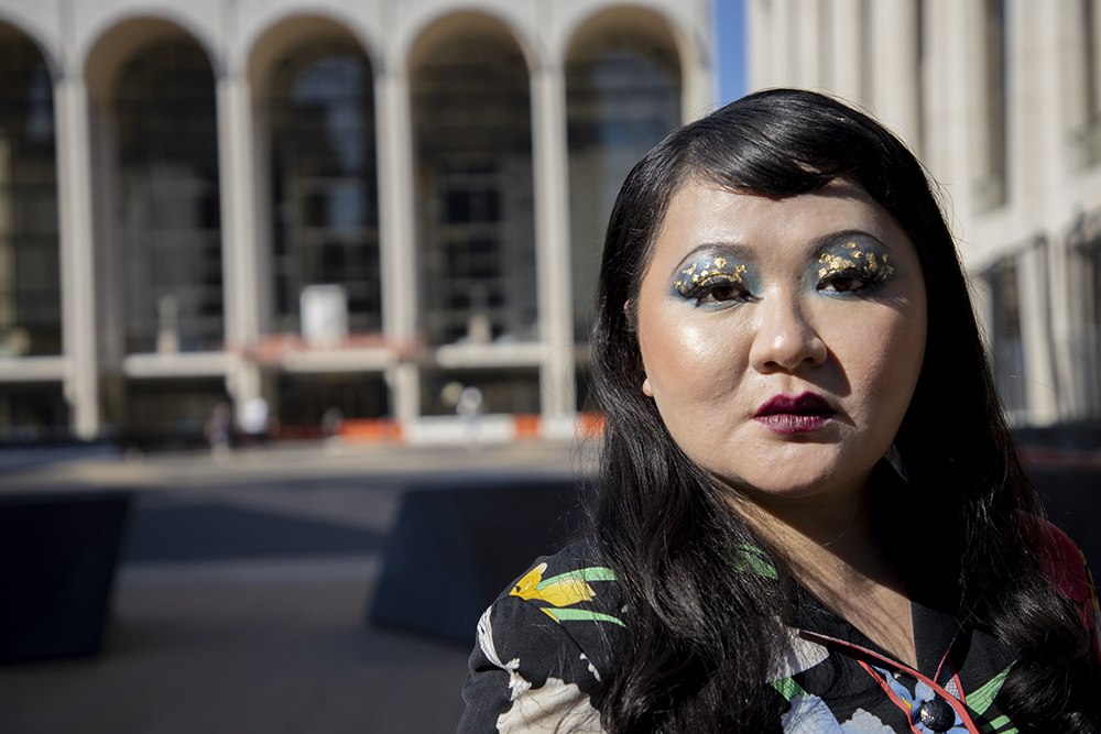 Du Yun Color photograph of an Asian woman wearing colorful makeup and glitter on her face as well as a floral print top; she stares at the camera in a serious manner. Behind her are the tall arches of the opera building at Lincoln Center in New York