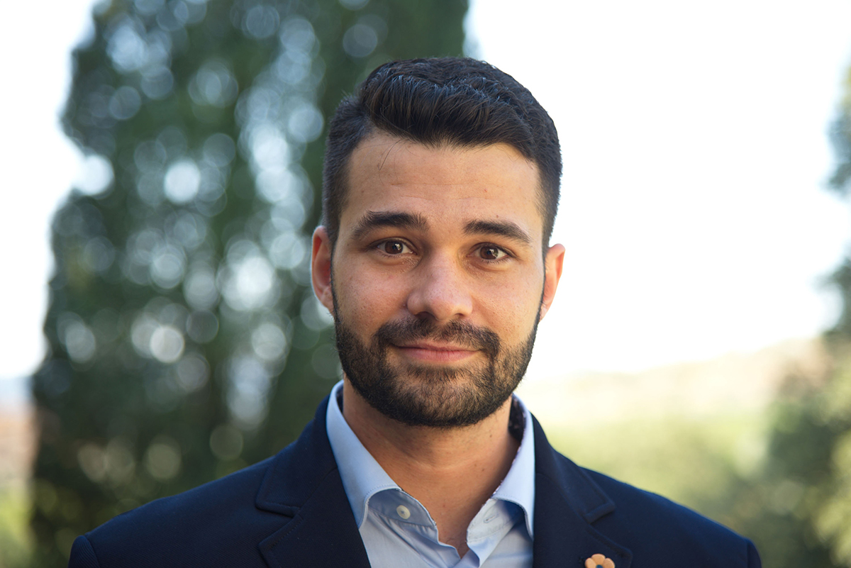 Edward Loss Color photograph of the head and shoulders of a light skinned man with dark hair and beard; he stands in an outdoor location with foliage behind him as he looks directly at the camera