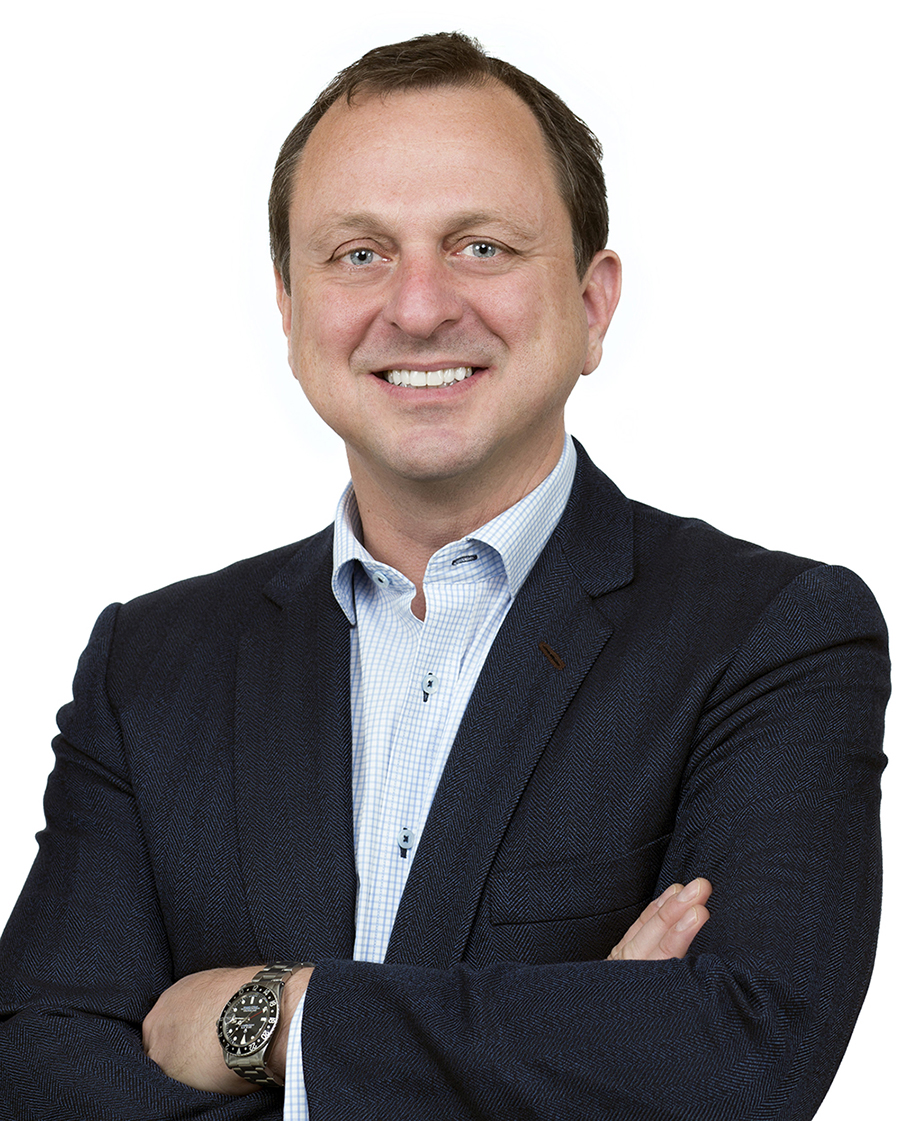 Color photograph of the head and torso of Gregg Pasquarelli wearing a blazer and standing with crossed arms against a white background