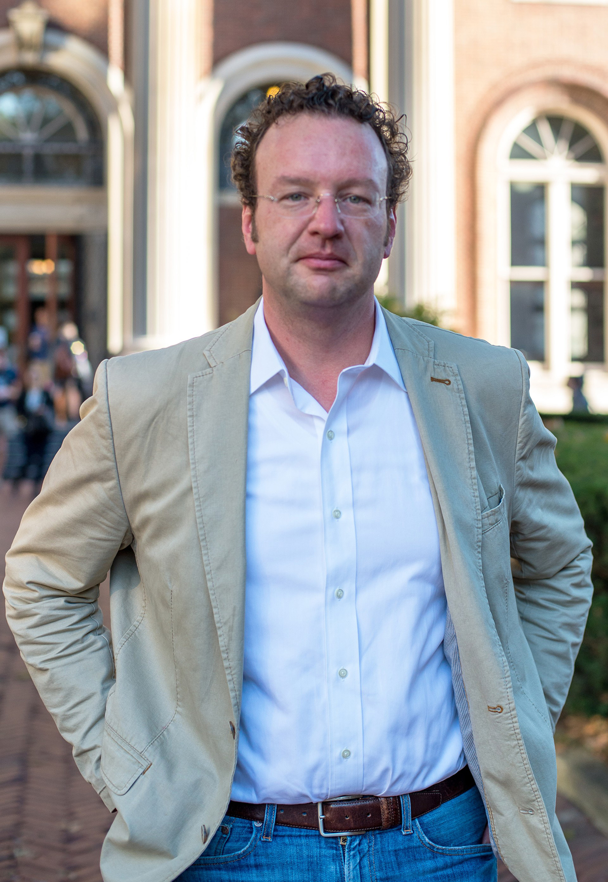 Color photo of a light skinned man wearing a tan blazer looking at the camera with hands on hips or in pockets