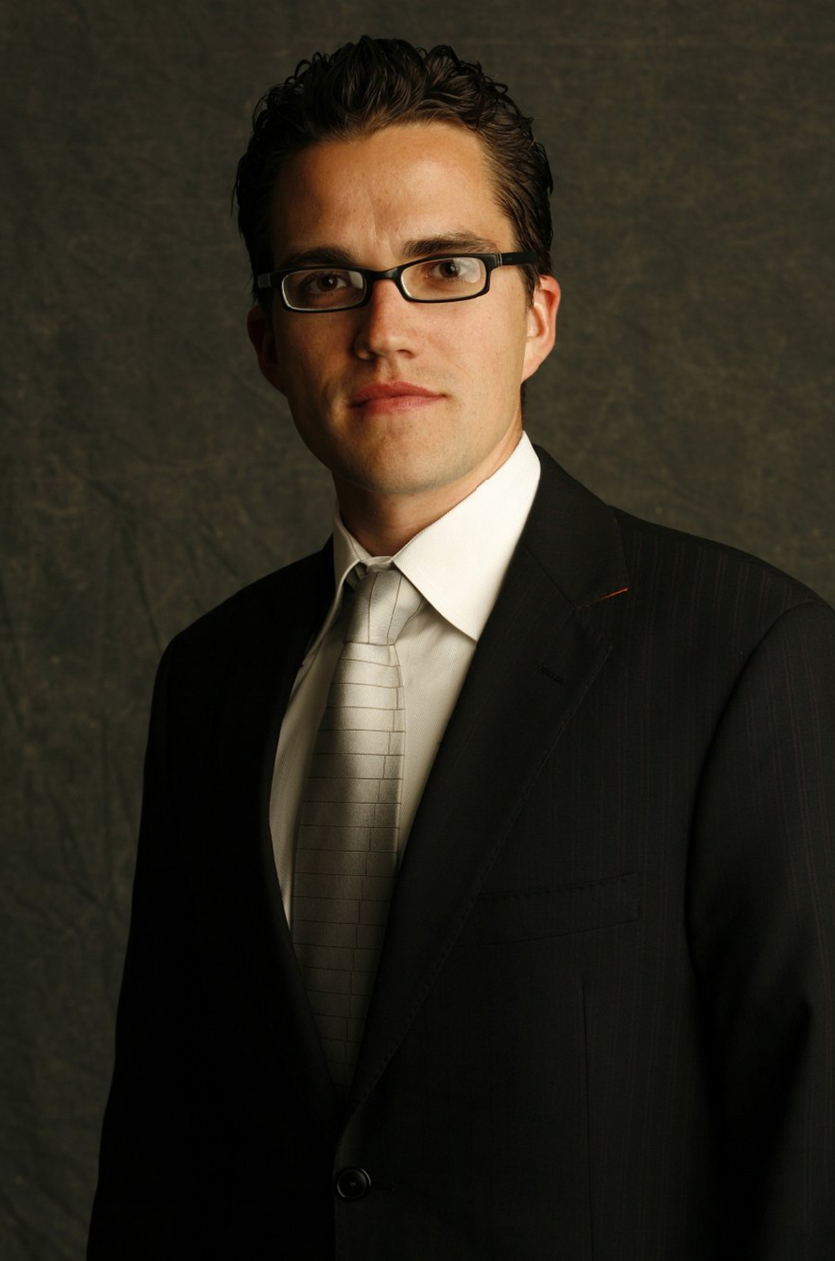 John Cary Color photograph of the head and torso of a light skinned man wearing glasses, dark suit, and tie, looking at the camera