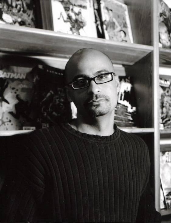 Junot Díaz Black and white photograph of an olive skinned man wearing a black shirt and glasses and standing in front of a shelf with comic books with the cover facing outward