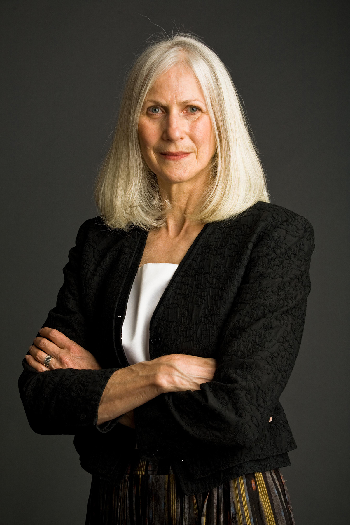 Color photograph of a light skinned woman from the waist up, standing in front of a photographer's studio backdrop and looking at the camera with her arms crossed
