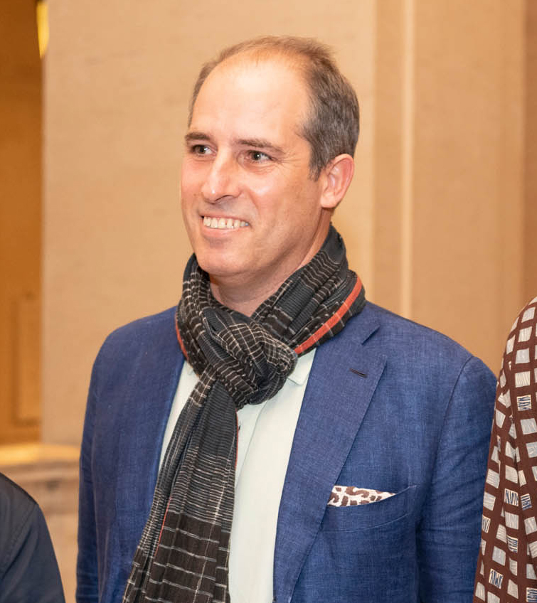 Color portrait of Peter Benson Miller from the waist up, wearing a blue suit jacket and striped scarf