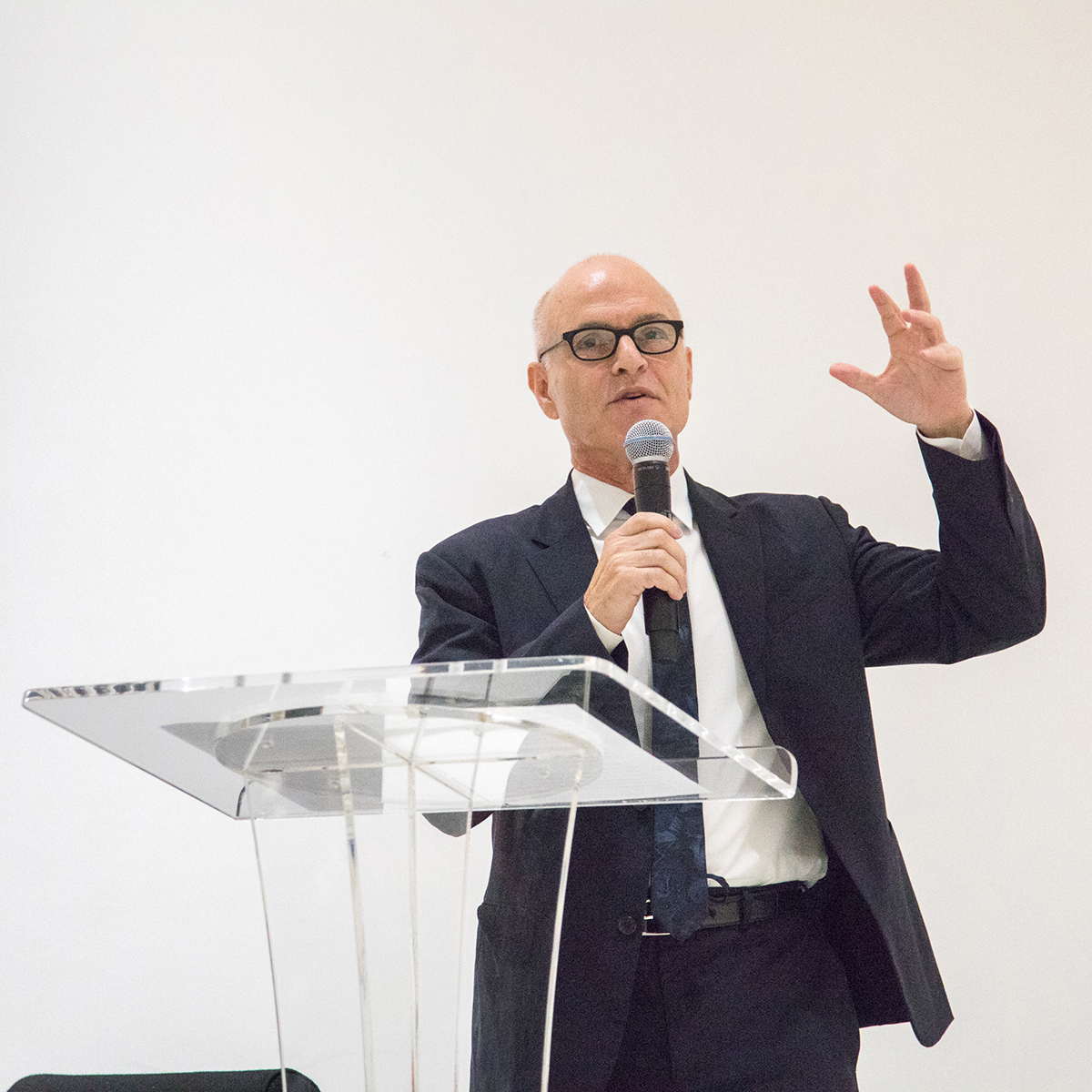 Staff - Mark Robbins Color photograph of a light skinned man wearing a dark suit and tie and standing at a transparent plastic podium; he gestures with his left hand as he holds and speaks into a cordless microphone in his right hand