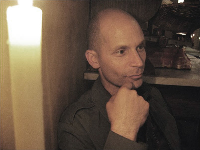 Tim Davis Color photograph of the head and shoulders of a light skinned man in an interior lit by a candle; one hand touches his face as he gazes off camera