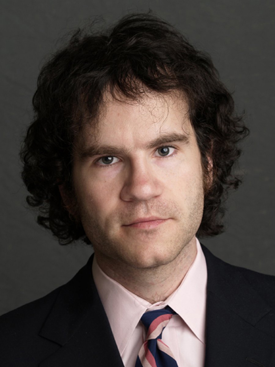 Tom Bissell Color photograph of the head of a light skinned man with brown hair looking at the camera without smiling