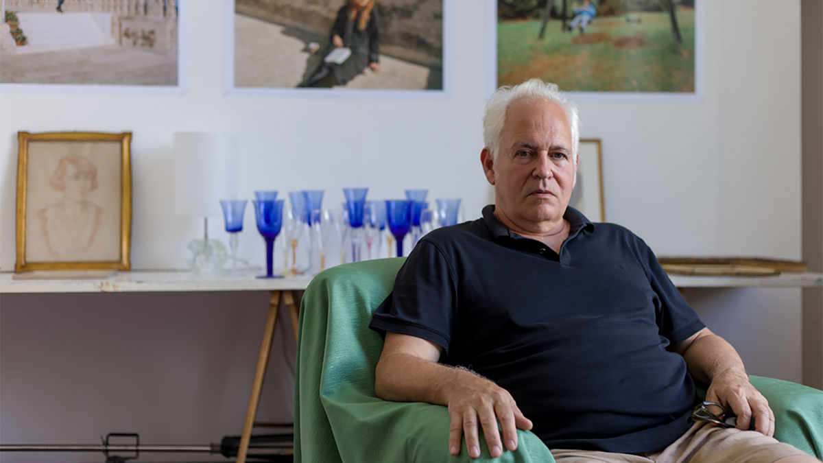 Color photograph of an older, light skinned man sitting in an armchair and looking at the camera