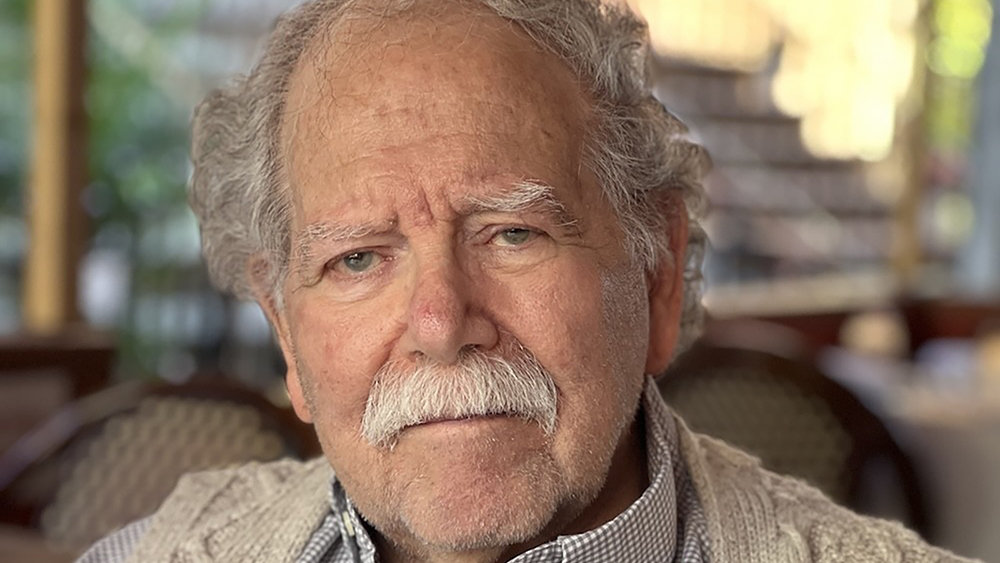 Color photo of the head and shoulders of an elderly light skinned man with a mustache looking at the camera with a serious expression