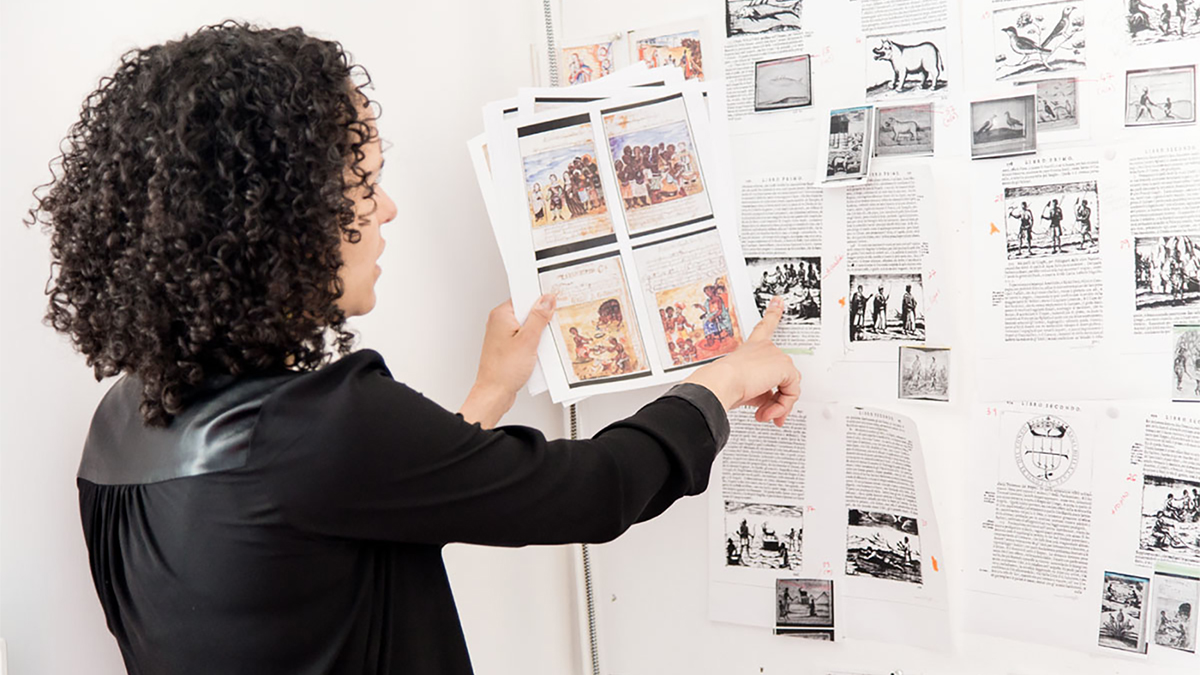 Color photo of the back of a woman pointing at a wall with many images pinned to it