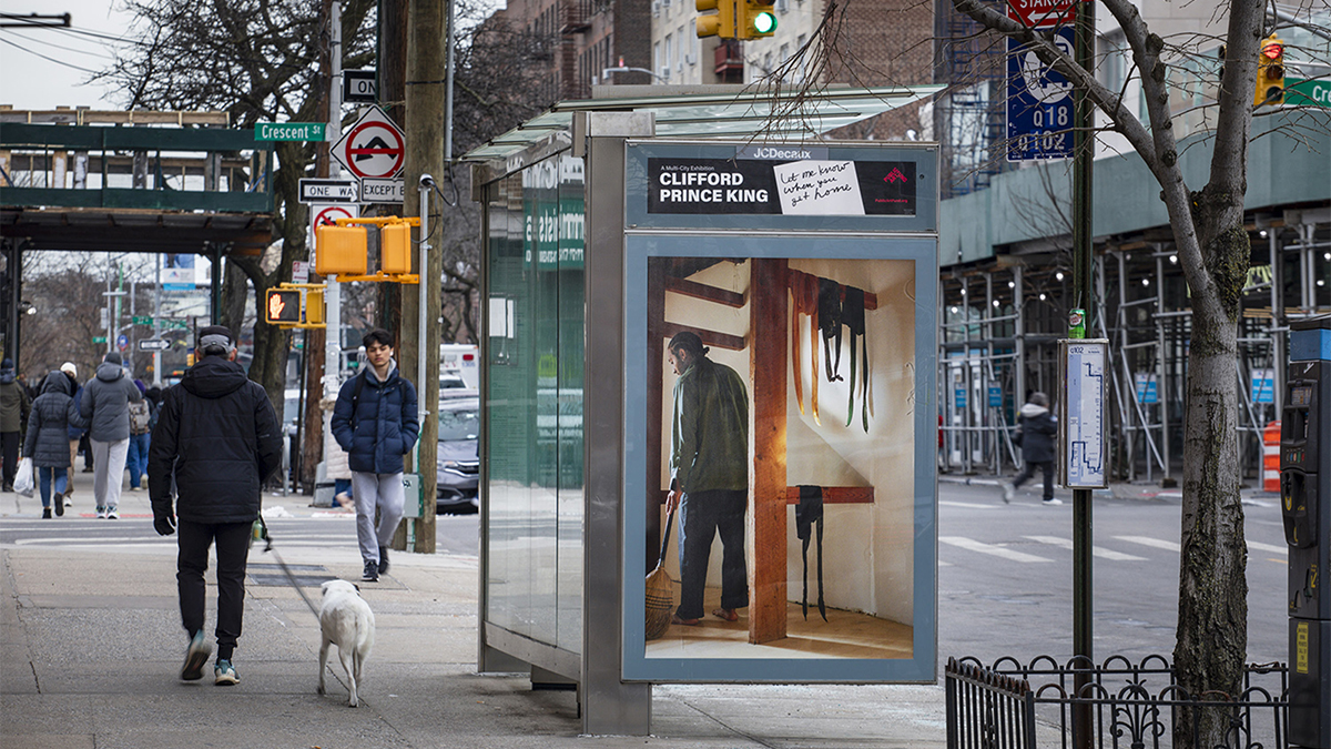 Color photo of a New York City street with a bus shelter than has a large artist's photo on the side