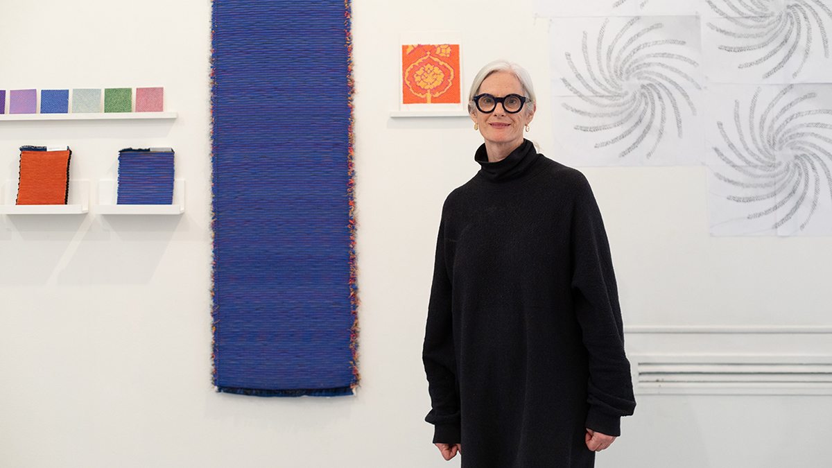 Color photo of an older light skinned woman wearing black and standing in front of her colorful fabric works fastened to a white studio wall