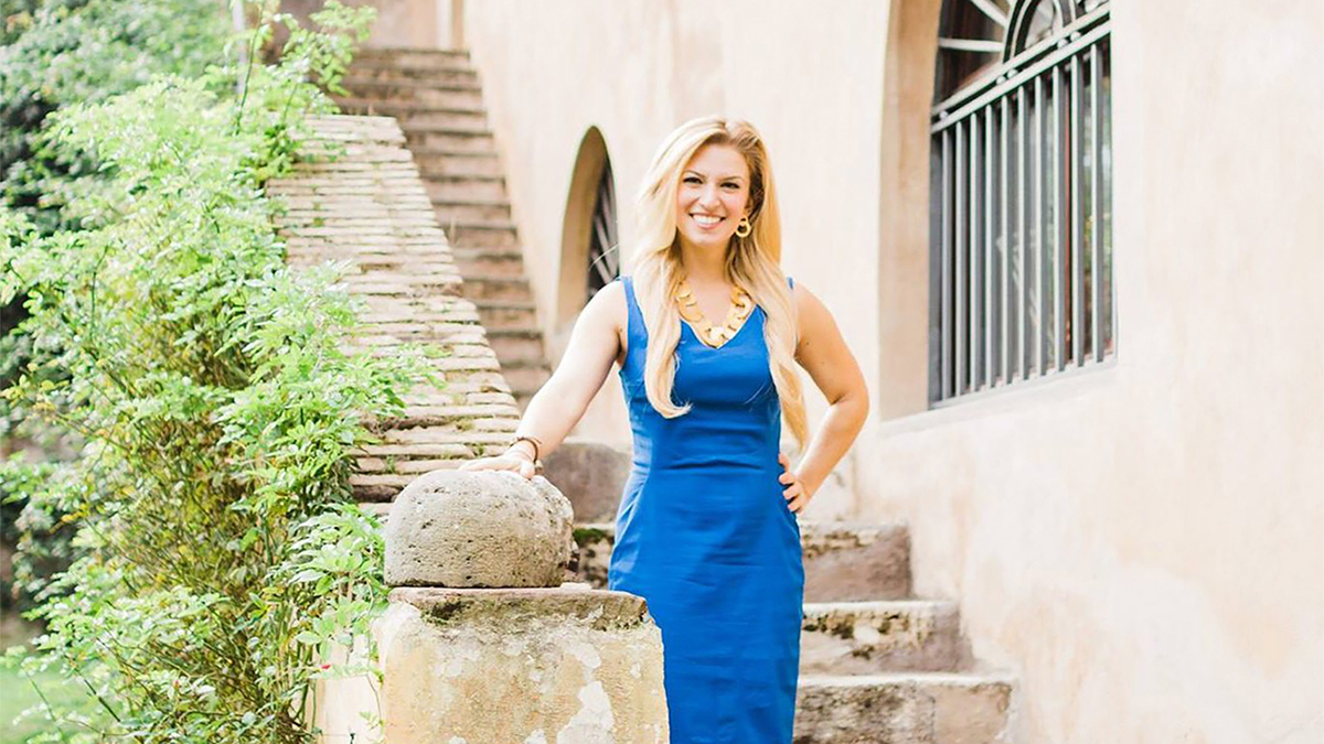 Color photograph of a light skinned woman with blond hair wearing a blue dress and smiling at the camera