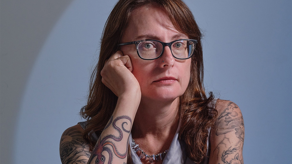Color photograph of a light skinned woman with tattoos and glasses looking at the camera