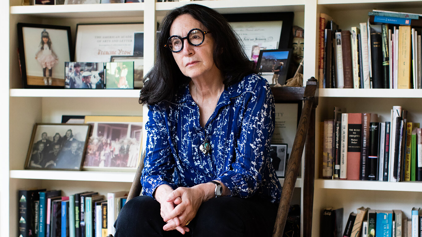 Color photograph of Francine Prose sitting on a stool in front of shelves of books and framed photos