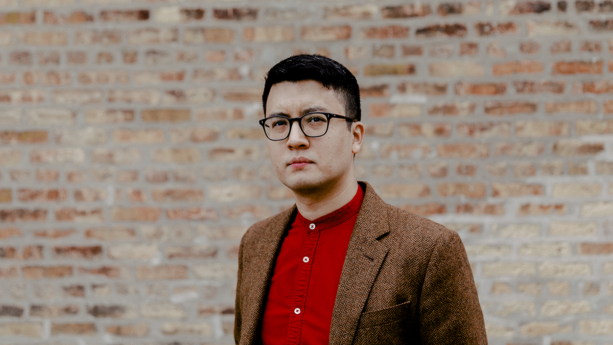 Color portrait of the torso of Igor Santo, wearing a red shirt, blazer, and glasses and standing in front of a brick wall