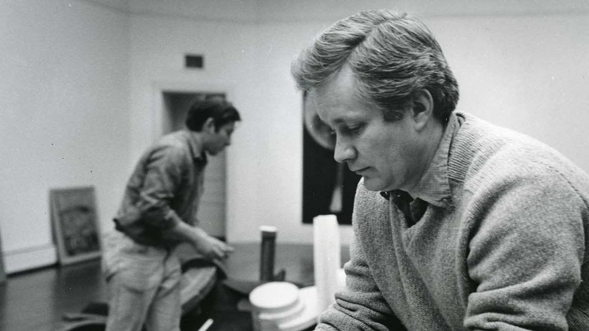 Black and white photograph taken in the late 20th century of a light skinned man wearing a sweater and looking toward the floor of an art gallery; behind him in the room is a second light skinned man