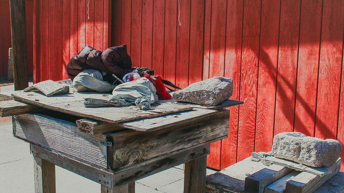Color photograph of stone and cloth objects on a makeshift wood table, outdoors in front of a red painted fence