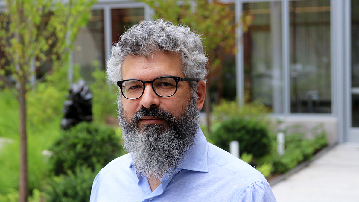 Color photo of the head and shoulders of a light skinned man with gray and black hair and bear, wearing glasses and looking at the camera