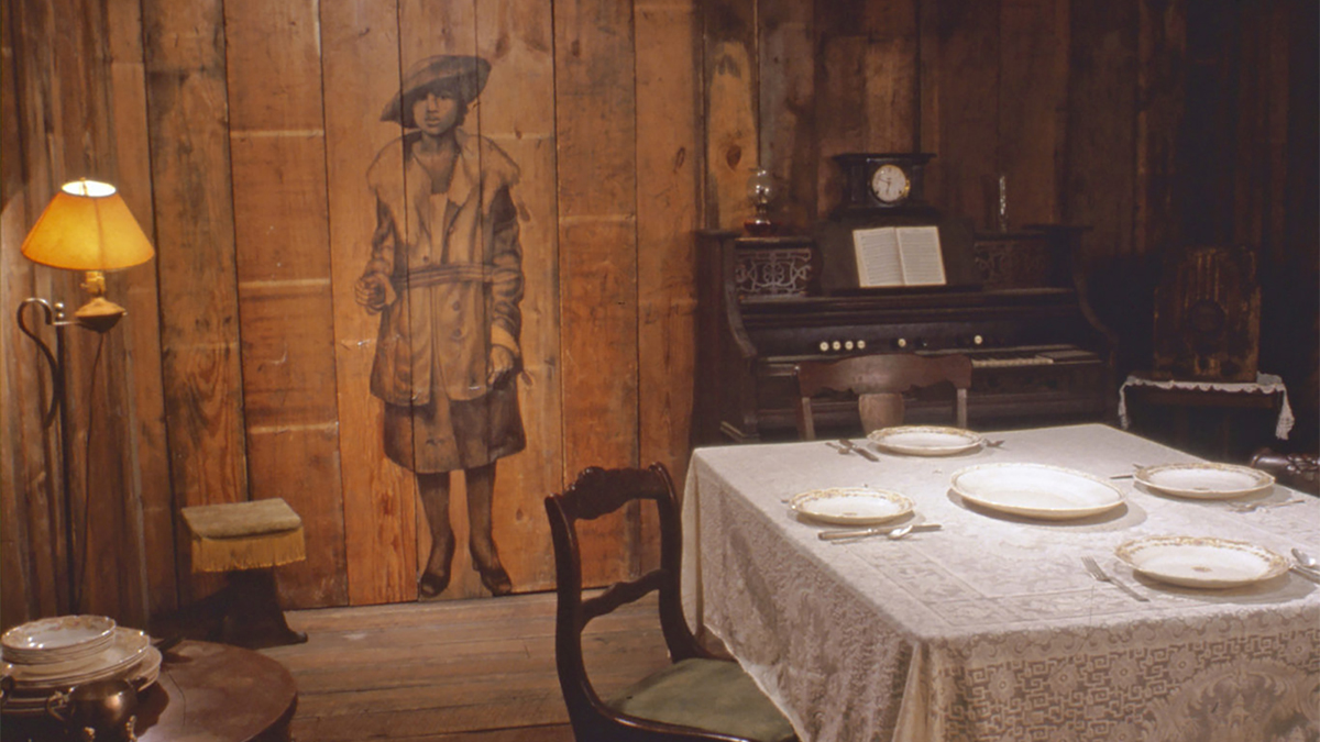 Color photo of an art installation with plates on a dining room table, a piano, and a drawing of a dark skinned woman directly on a wood wall