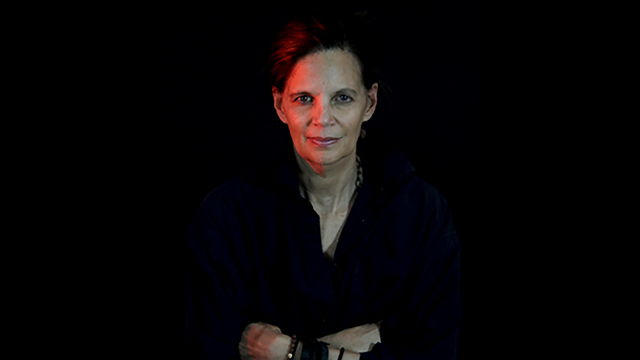 Color photograph of the head and torso of a woman looking straight at the camera with her arms folded against her chest