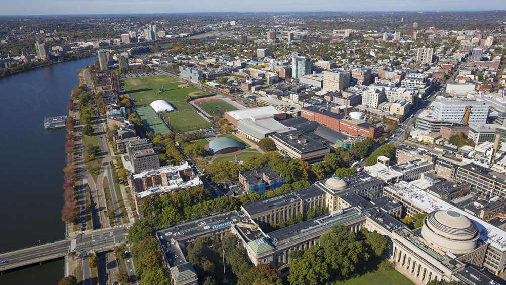 Aerial color photograph of the MIT campus in Cambridge, Massachusetts, showing numerous neoclassical and modern buildings, the Charles River to the left, and various sports fields