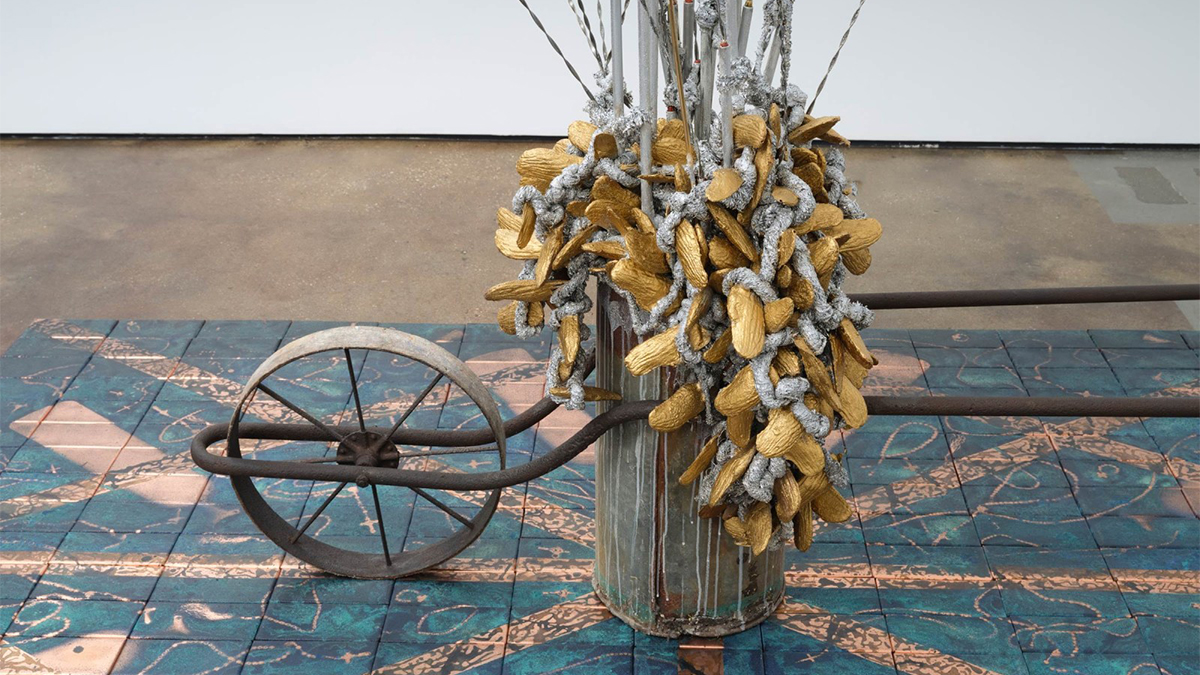 Color photograph of a detail of a contemporary sculpture with a wheel and flower stems resting on painted ceramic tiles