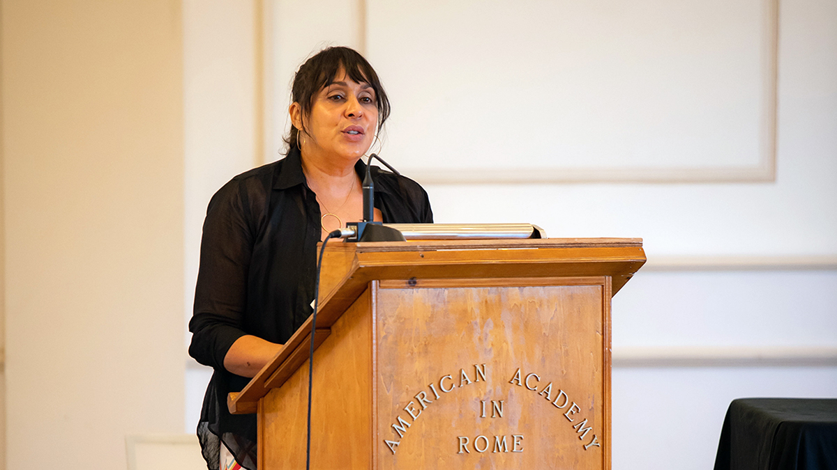 Color photograph of a brown skinned woman with dark hair standing at a podium in a lecture room and speaking